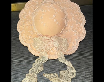 May include: A peach-colored bonnet with a scalloped brim and a matching bow. The bonnet is adorned with white floral embroidery and small pearl accents. The lace bow has long, flowing ribbons.