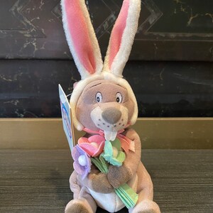 Könnte beinhalten: Ein Stofftier-Hase mit langen, rosa gefütterten Ohren und einem weißen Hut. Der Hase hält einen Strauß pastellfarbener Blumen und hat ein rosa Band um den Hals. Das Spielzeug hat ein Etikett.