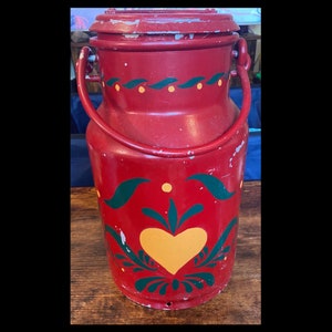 Pode incluir: Uma lata de leite vermelha vintage com tampa e alça. É decorada com desenhos florais verdes e amarelos, incluindo um coração amarelo. A lata mostra sinais de desgaste, aumentando seu charme antigo.