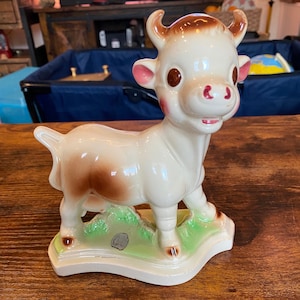 May include: A ceramic cow figurine featuring a cream-colored body with brown patches, brown horns, and pink-tinted ears and cheeks. The cow stands on a green base with a small gray circular mark on the front, displaying large brown eyes and a smile.