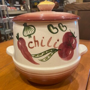 Op de afbeelding: Een crèmekleurige keramische chilipot met bijpassend deksel, voorzien van handgeschilderde afbeeldingen van rode tomaten, een wortel, sperziebonen en een ui. Het woord chili staat in bruine letters op de pot en het glazuur vertoont barstjes.