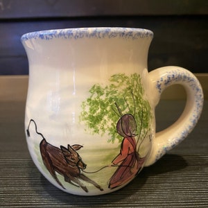 May include: White ceramic mug with a blue speckled rim and handle. The mug features a hand-painted scene of a person in a red dress leading a brown animal on a leash. Green foliage is painted in the background.