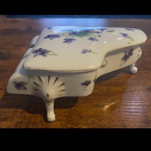 Può includere: Un portagioie a forma di pianoforte in porcellana bianca con coperchio incernierato. La scatola è decorata con motivi floreali viola e finiture dorate. Il pianoforte ha tre piccole gambe e un pannello frontale decorativo.