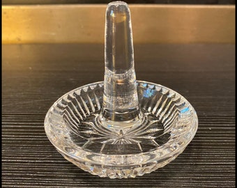 May include: A clear glass ring holder with a round base and a tall, cylindrical post. The base features a starburst design and a textured, scalloped edge. The ring holder is designed to display rings and other small jewelry items.