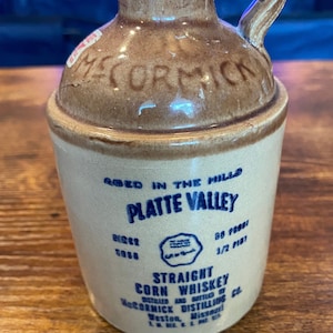 May include: A vintage ceramic jug of Platte Valley Straight Corn Whiskey. The jug is tan and brown with a handle and embossed text. The label reads "Aged in the Hills" and "McCormick Distilling Co."