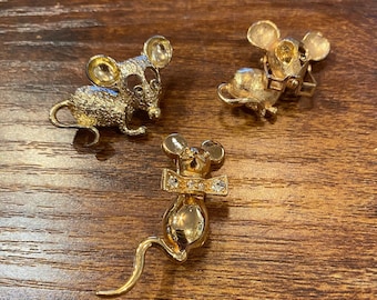 May include: Three gold-toned mouse brooches. One mouse has a textured body, another appears to be reading, and the third has a rhinestone bow tie. The brooches are displayed on a wooden surface.