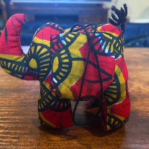May include: A stuffed elephant toy crafted from fabric featuring a vibrant pattern of red, yellow, and black geometric shapes. The elephant has a raised trunk, a small black yarn tail, and a thin black string loop attached to its back for hanging.