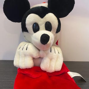 May include: A plush Mickey Mouse toy with black ears, a white face, and a red outfit. The toy has black eyes, a black nose, and white gloves with black lines. The red outfit is visible at the bottom of the image.