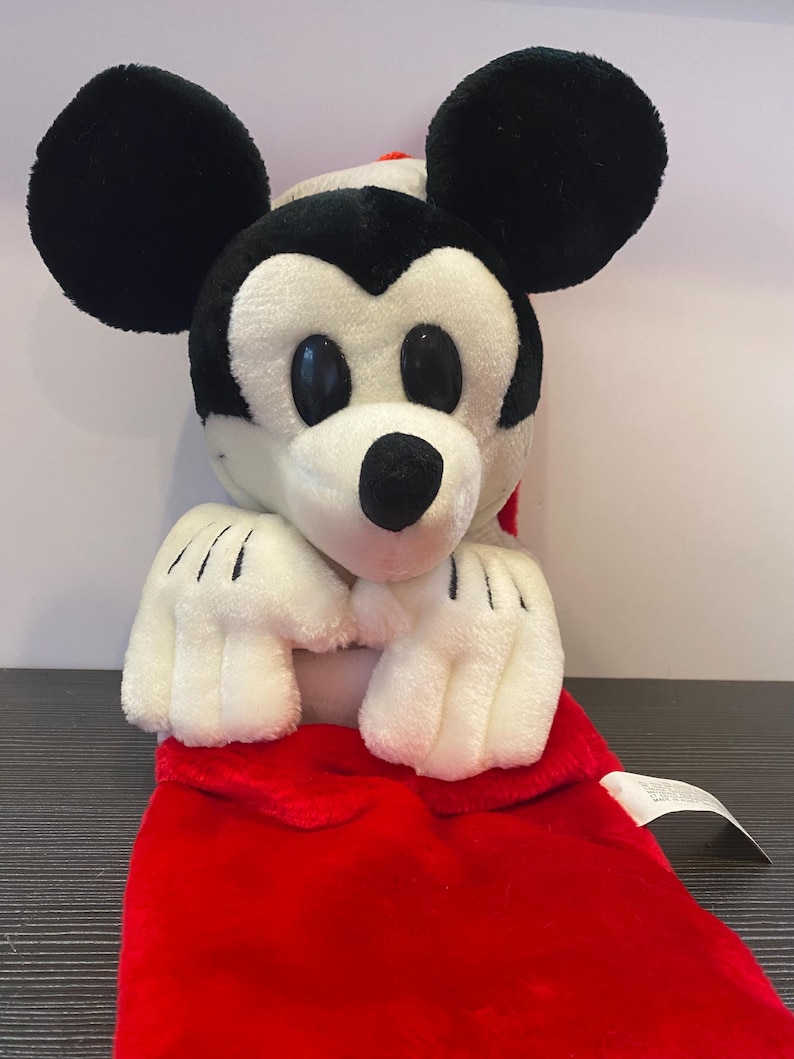 May include: A plush Mickey Mouse toy with black ears, a white face, and a red outfit. The toy has black eyes, a black nose, and white gloves with black lines. The red outfit is visible at the bottom of the image.