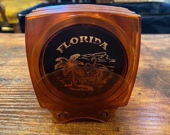Puede incluir: Un portavasos vintage de color ámbar con un emblema circular negro. El emblema presenta la palabra "FLORIDA" en letras doradas, junto con una palmera y un flamenco. El soporte está hecho de un material plástico transparente.