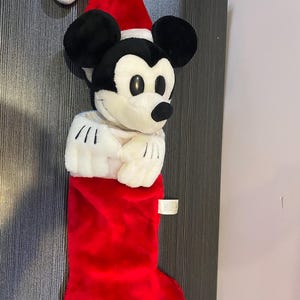 May include: A plush Christmas stocking featuring Mickey Mouse wearing a Santa hat. The stocking is red with a white cuff, and Mickey's face and hands are visible. A festive holiday decoration.