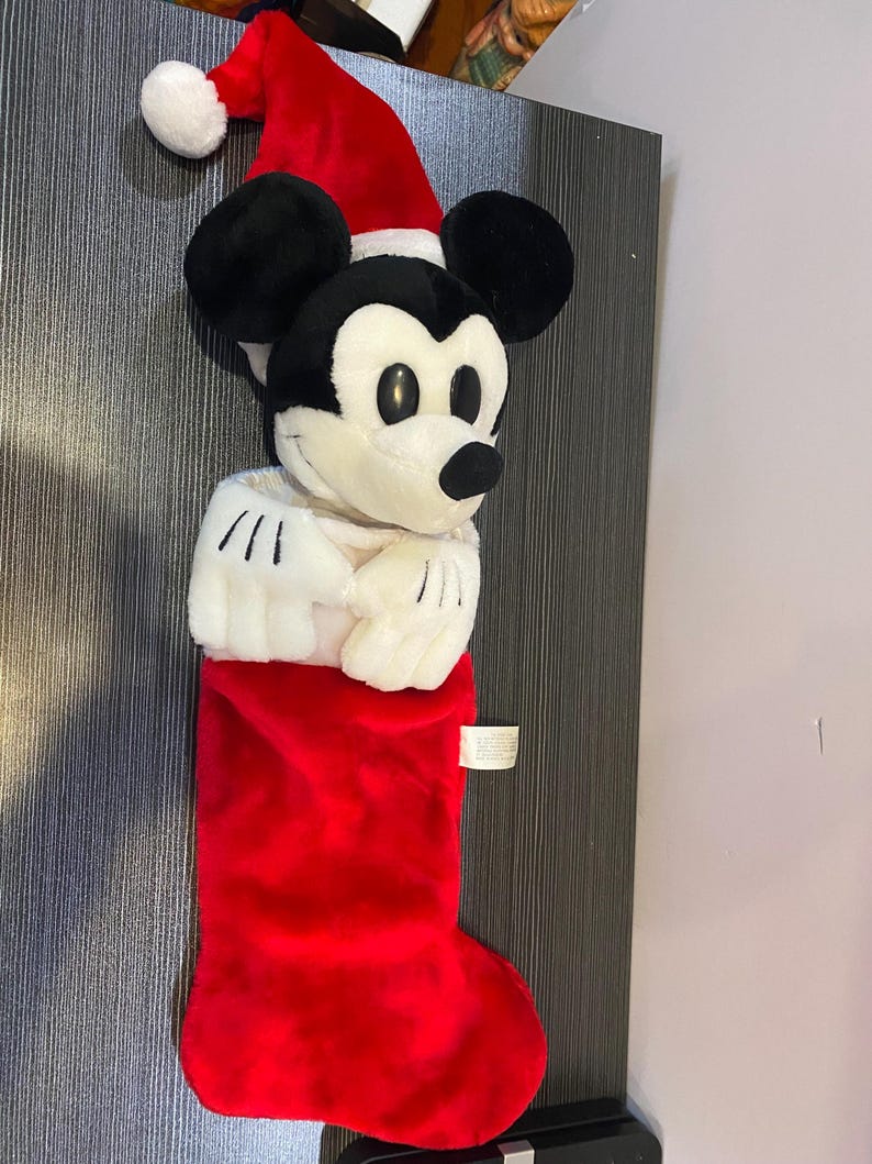 May include: A plush Christmas stocking featuring Mickey Mouse wearing a Santa hat. The stocking is red with a white cuff, and Mickey's face and hands are visible. A festive holiday decoration.
