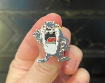 May include: Enamel pin featuring the Tasmanian Devil cartoon character. The pin has a gray and white body with a red tongue and eyes. The pin is held in a person's hand.