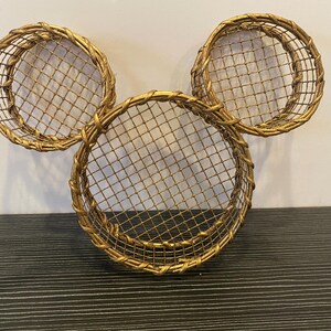 May include: Three gold-colored wire baskets arranged in a Mickey Mouse head shape. The baskets have a woven border and a diamond-patterned wire mesh. The design is a decorative wall hanging or storage solution.