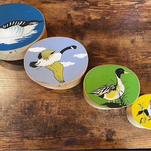 May include: Four round wooden boxes with painted duck illustrations. The lids feature a variety of duck designs, including a duck swimming on blue water, a duck in flight, and ducks in a grassy area. The boxes are of varying sizes.