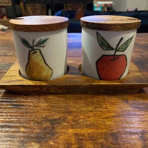 Puede incluir: Un juego de dos tarros de cerámica blanca con tapas de madera, cada uno decorado con un diseño de fruta pintado a mano. Uno presenta una pera amarilla, el otro una manzana roja. Los tarros están sobre una bandeja de madera.
