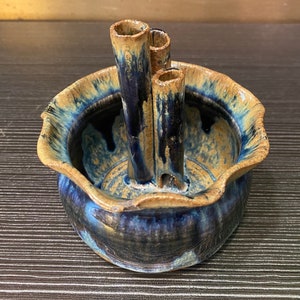 May include: A ceramic vase with a distinctive design. The vase has a round base with a wavy rim and three vertical tubes. The colour scheme includes shades of blue, brown, and tan, creating a rustic aesthetic. Ideal for displaying flowers.