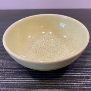 May include: A small, round ceramic bowl with a pale green interior and a speckled pattern. The bowl has a smooth, slightly raised rim and a glossy finish. The exterior is a solid, light green color.