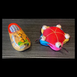 Puede incluir: Dos objetos decorativos sobre una superficie oscura. Uno es un zueco de madera con un cojín a cuadros azul y blanco en el interior y detalles pintados. El otro es un alfiletero rojo, azul, amarillo y rosa con seis pequeñas cabezas de muñeca.