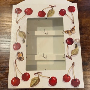 Könnte beinhalten: Weißer Schlüsselhalter aus Holz mit rechteckiger Öffnung und gewölbter Oberseite. Der Schlüsselhalter ist mit roten Kirschen und grünen Blättern verziert. Zwei Schmetterlinge sind ebenfalls auf die Vorderseite gemalt. Der Schlüsselhalter hat mehrere Haken im Inneren.