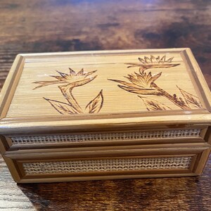 May include: A rectangular bamboo box with a hinged lid. The lid features two etched bird of paradise flowers. The box has a woven texture around the middle and is a light brown colour.