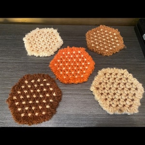 Könnte beinhalten: Fünf handgefertigte, sechseckige Häkeluntersetzer in verschiedenen Farben. Die Untersetzer umfassen Beige-, Braun- und Orangetöne. Jeder Untersetzer hat ein Sternmuster in einer Kontrastfarbe, was dem Set einen optischen Reiz verleiht.