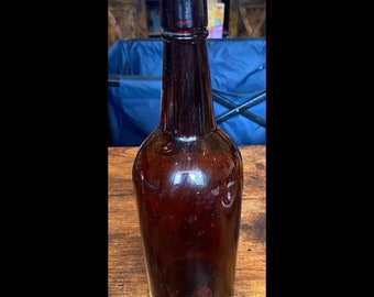 Puede incluir: Una botella de vidrio alta y vacía de color marrón ámbar profundo. La botella presenta un cuerpo cilíndrico, un cuello delgado y un borde grueso y redondeado en la abertura superior. La superficie de vidrio es lisa y refleja la luz.