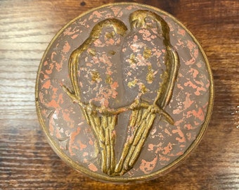 May include: Round, decorative box with a bird motif. The lid features two birds perched on a branch, rendered in gold-colored metal. The background has a speckled, pinkish-brown pattern. The box has a gold-colored rim.