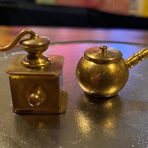 Op de afbeelding: Twee kleine, goudkleurige items: een koffiemolen en een potje met een deksel. De molen heeft een slinger en een vierkante basis. De pot heeft een afgeronde body en een lange handgreep. Beide zijn gemaakt van glanzend metaal.