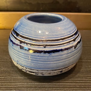 May include: A small, round ceramic vase with a light blue base colour. The vase features horizontal bands of black and white stripes. The opening is at the top. The vase is suitable for home decor.