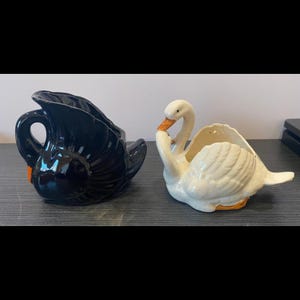 Könnte beinhalten: Zwei Schwanfiguren aus Keramik. Eine ist schwarz mit einem orangefarbenen Schnabel, die andere ist weiß mit einem orangefarbenen Schnabel. Beide haben ein dekoratives Design und stehen auf einer dunklen Oberfläche. Die Schwanfiguren sind wahrscheinlich Dekorationsartikel.