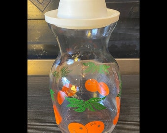 May include: A clear glass pitcher with a white plastic lid. The pitcher is decorated with orange fruit and green leaves. The pitcher is empty and sitting on a dark surface.