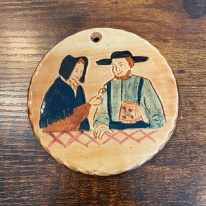 May include: Round ceramic wall hanging with a painted scene of two figures. One figure wears a black hat and holds a mug, the other wears a dark blue shawl. The piece has a hole for hanging.