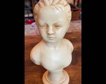 May include: A cream-colored bust of a young person with a blue bow in their hair. The sculpture has detailed facial features and is mounted on a pedestal base. The bust is likely made of ceramic or a similar material.