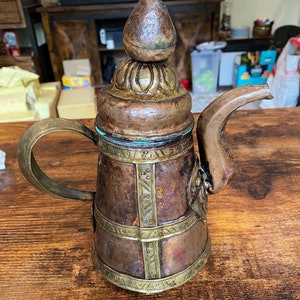 May include: An antique copper and brass teapot with a decorative handle and spout. The teapot features ornate detailing, including a textured lid and bands of brass around the body. The overall color palette is a mix of brown, copper, and gold.