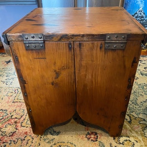 K&ouml;nnte beinhalten: Ein kleiner, rustikaler Holzschrank mit Klappdeckel und zwei T&uuml;ren. Der Schrank ist aus hellbraunem Holz mit sichtbarer Holzmaserung und Metallscharnieren gefertigt. Die Ecken sind mit Metallwinkeln verst&auml;rkt. Der Schrank hat ein gealtertes Aussehen.