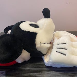 May include: A plush Mickey Mouse head and glove. The Mickey Mouse head is black and white with a black ear and a black nose. The glove is white with black lines. The plush toy is on a black surface.