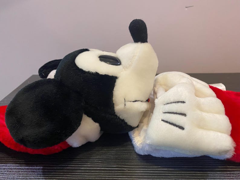 May include: A plush Mickey Mouse head and glove. The Mickey Mouse head is black and white with a black ear and a black nose. The glove is white with black lines. The plush toy is on a black surface.