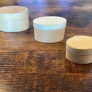 May include: Three small, round, wooden boxes with lids are arranged on a dark brown wooden surface. The boxes are light beige with a natural wood grain. One box has a white band around the middle.