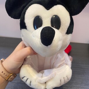 May include: A plush Mickey Mouse toy with black ears, a black nose, and large black oval eyes. The toy is primarily white with a red accent and is held in a person's hand.