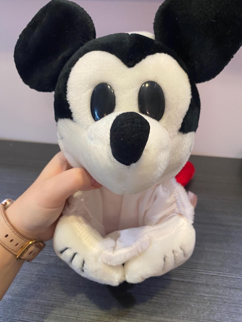 May include: A plush Mickey Mouse toy with black ears, a black nose, and large black oval eyes. The toy is primarily white with a red accent and is held in a person's hand.