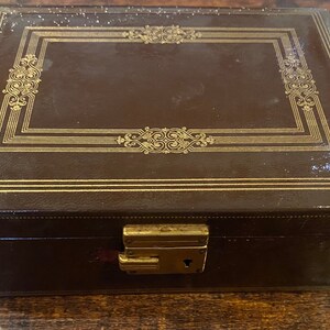 May include: A rectangular, brown leather box with gold-colored decorative trim and a clasp. The box features a rectangular design on the lid with ornate gold detailing. The box is closed and appears to be vintage.
