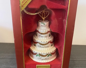 May include: A white, tiered Christmas ornament shaped like a cake, with red ribbon and gold trim. The ornament is in a red box with the text "2007 Annual" and "Our 1st Christmas Together". The Lenox logo is visible.