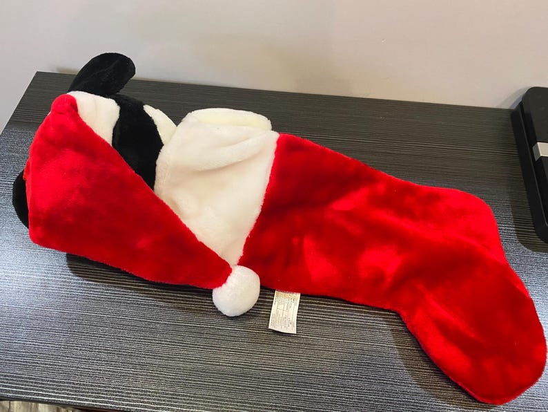 May include: A plush Christmas stocking in the shape of a character's head, featuring a red Santa hat with a white trim and a white pom-pom. The stocking is primarily red with a white cuff.
