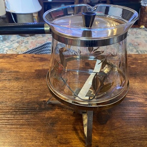 Könnte beinhalten: Eine Vintage-Kaffeekanne aus Glas mit schwarzem Griff und Deckel. Die klare Glaskanne hat ein dekoratives Blättermuster und steht auf einem Metallständer. Die Kanne hat ein silberfarbenes Metallband oben und unten.