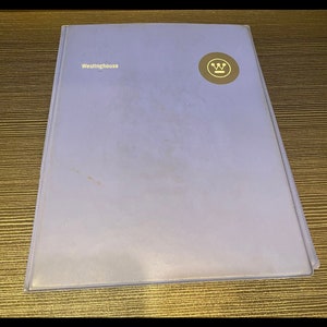 May include: A light purple binder with the word "Westinghouse" printed in gold on the front cover. A gold-colored circular logo is on the upper right corner of the binder. The binder is rectangular and appears to be made of a smooth material.