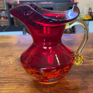 Puede incluir: Una jarra de vidrio rojo rubí con un asa transparente y un acento dorado decorativo en la base. La jarra tiene un pico acampanado y un cuerpo bulboso. El vidrio tiene un acabado brillante y refleja la luz.