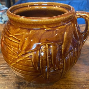 May include: A brown ceramic pot with a handle. The pot has a rounded shape with a textured surface and a glossy finish. The handle is attached to the side of the pot. The pot is sitting on a wooden surface.