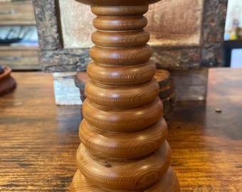 Puede incluir: Un candelabro de madera de color marrón medio con un diseño apilado y escalonado, que presenta una base circular ancha, una serie de anillos redondeados graduados que forman la columna central y una plataforma superior plana con una hendidura.