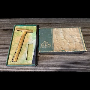 Puede incluir: Una maquinilla de afeitar de seguridad dorada vintage en su caja original verde y crema. La maquinilla está en una caja verde abierta con un interior crema. La caja de al lado es verde con las palabras "New GEM Micromatic" impresas.